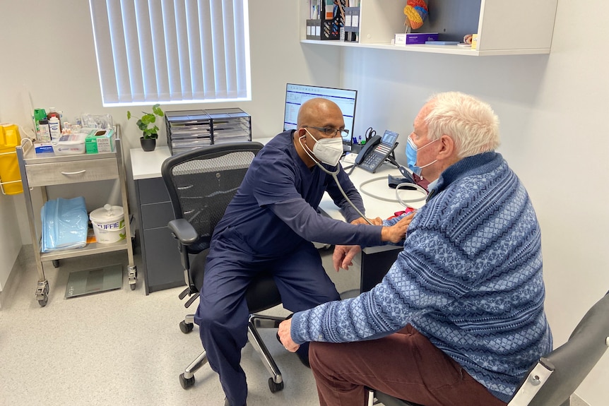 A doctor listens to a patient's heart with a stethoscope while seated at an office desk