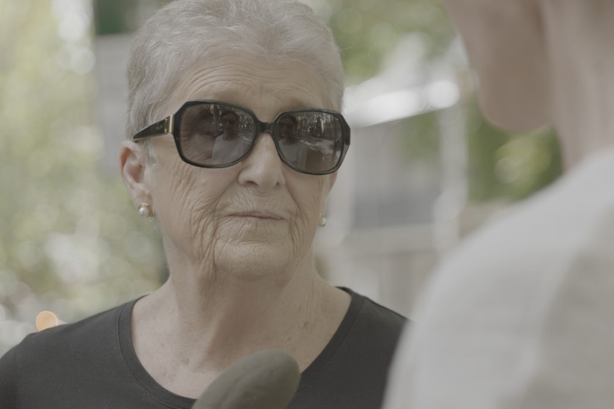 A woman with grey hair, black shirt and sunglasses looking towards someone interviewing her with a microphone.