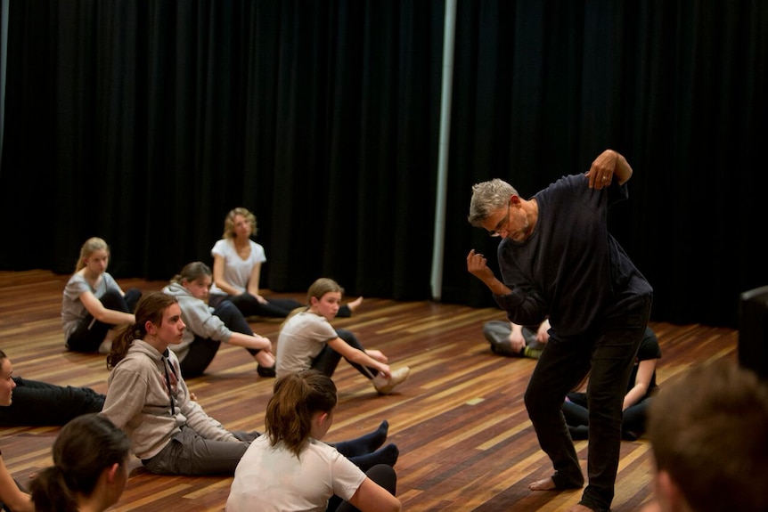 A man, standing but bent, showing a dance move to several young people sitting on a smooth studio floor.