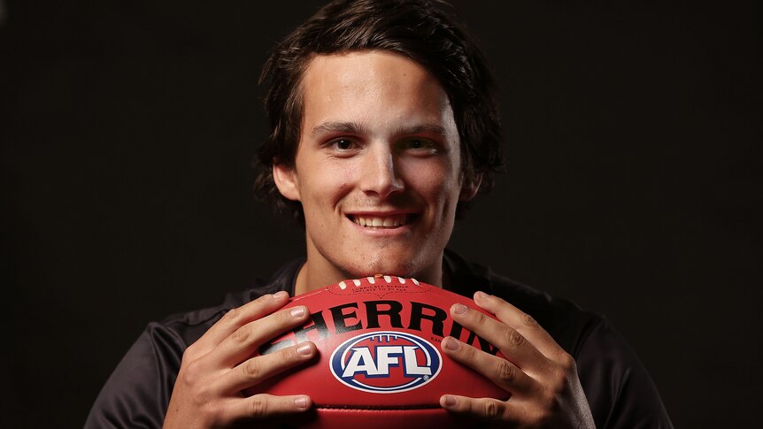 AFL player Harley Balic holds a football in two hands while smiling at the camera.