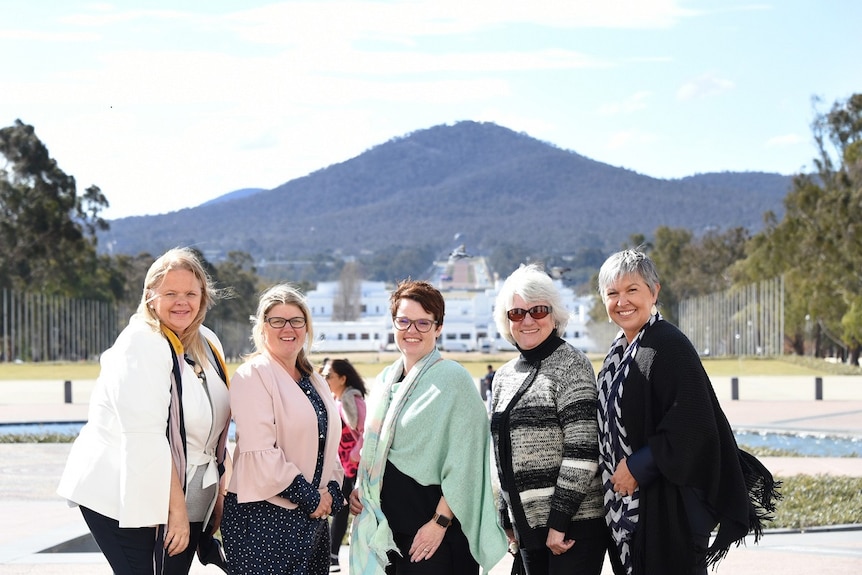 A woman in a white jacket and four more women stand smiling in with blue sky and a huge mountain in the background.