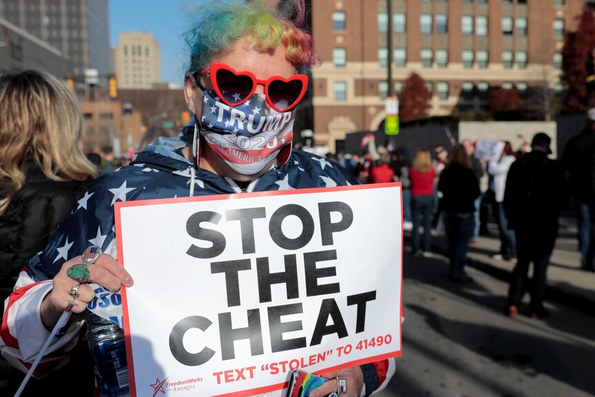 A woman in heart sunglasses with rainbow hair and a Trump face mask holds a placard that reads Stop the Cheat