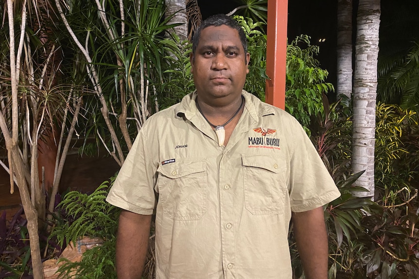 A man dressed in khaki and wearing a necklace stands in front of tropical trees at night