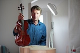 Peter Goodfellow stands at his work bench holding a violin.
