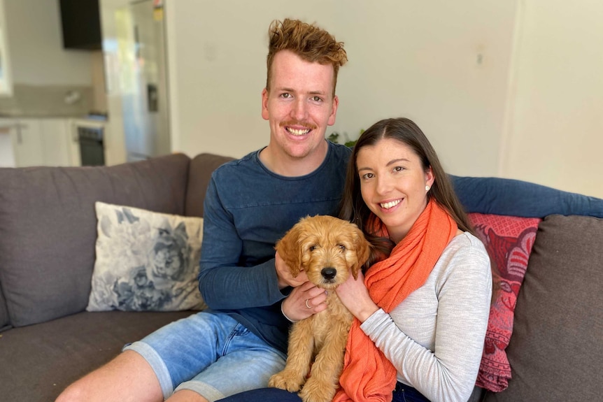 A man and a woman sitting on a sofa holding a light brown puppy