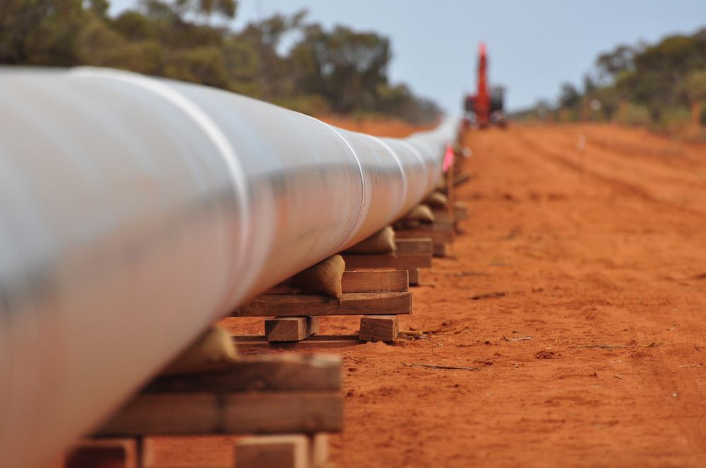 A section of metal pipeline stretching into the distance lies in wooden blocks on the ground, with a crane in the background.