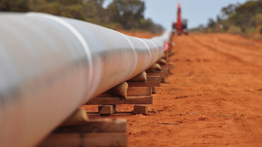 A section of metal pipeline stretching into the distance lies in wooden blocks on the ground, with a crane in the background.