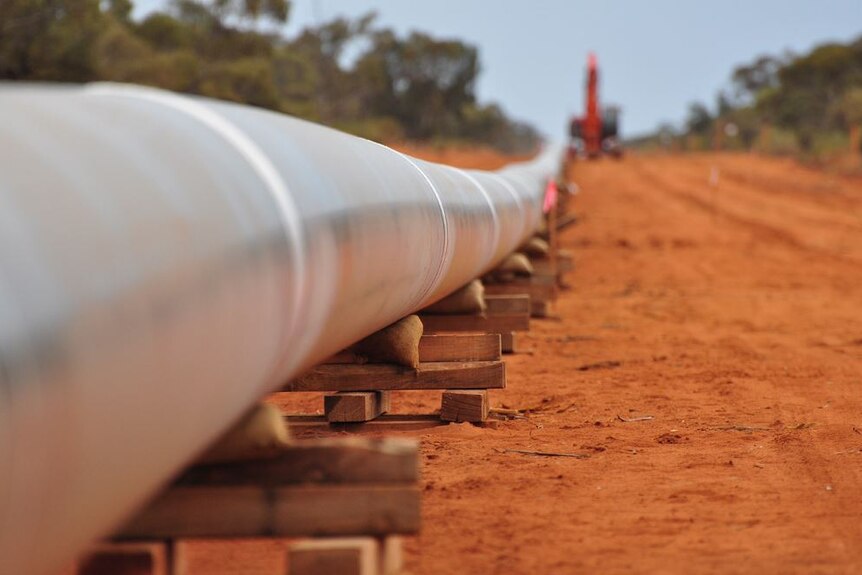 A section of metal pipeline stretching into the distance lies in wooden blocks on the ground, with a crane in the background.
