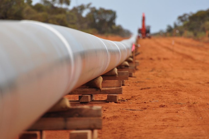 Une section de pipeline métallique s'étendant au loin repose sur des blocs de bois au sol, avec une grue en arrière-plan.