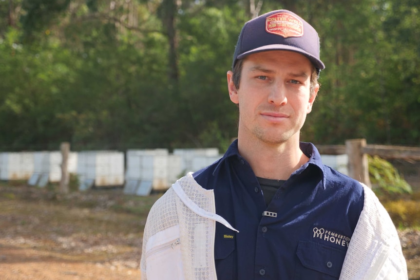 A man in a baseball cap and shirt that says 'Pemberton Honey Company' stands against the backdrop of a forest.