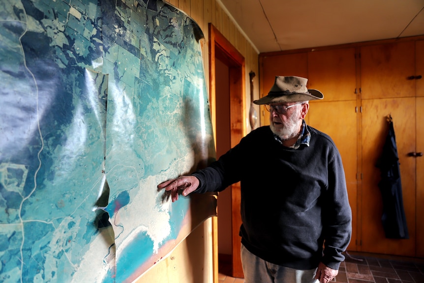 Older man with a weathered hat points at large photographic aerial map inside a wood paneled room