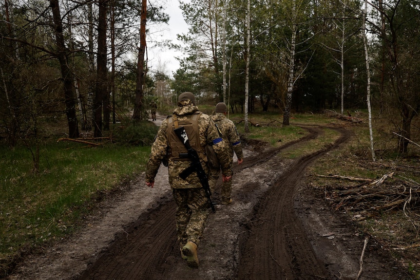 Two soldiers wearing camouflage gear including heavy coats, one carrying a gun, walk down a muddy path through a forest