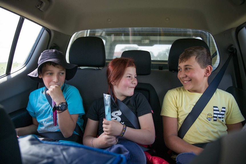 Three students laugh while sucking on ice blocks in the back seat of a car.