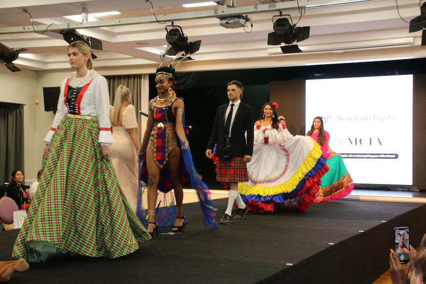 Models from diverse backgrounds and dressed in unique outfits walk along a catwalk at a fashion show.