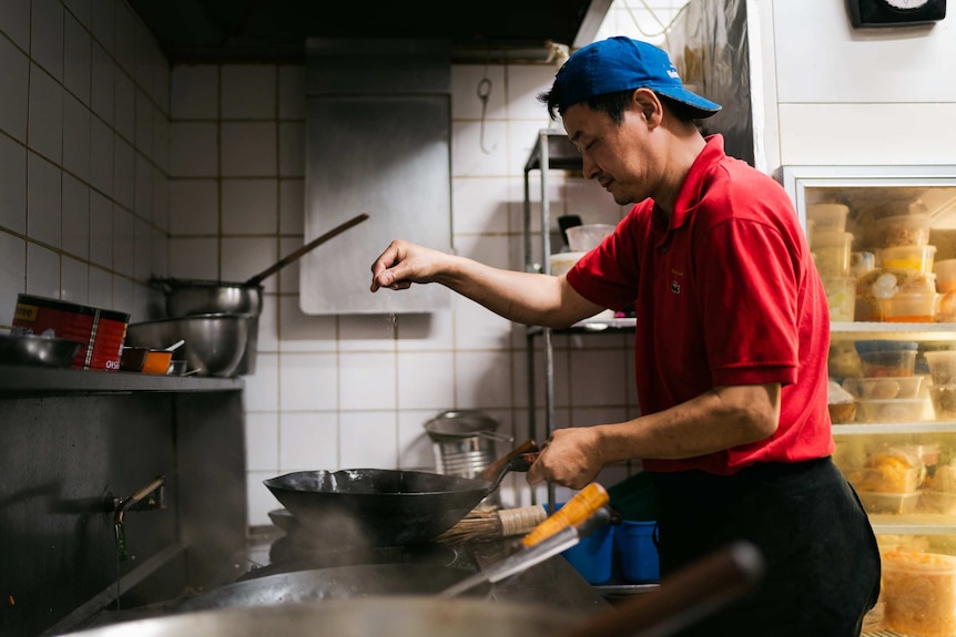 A chef wearing a red shirt and blue hat adds salt to a wok in a kitchen