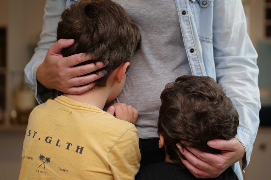 A mother hugs two young boys to her waist.
