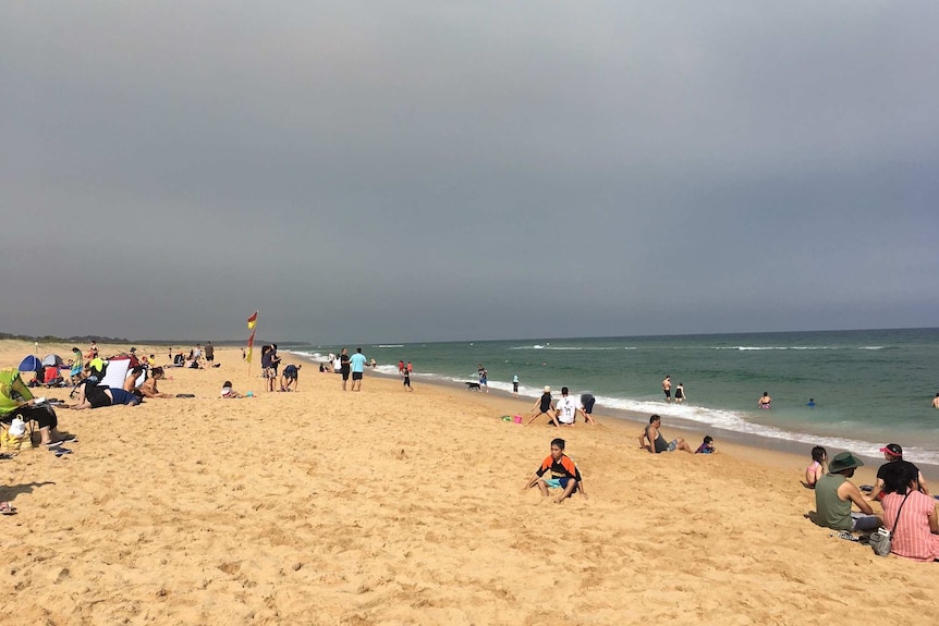 Families sit on a beach next to a calm ocean with a dark grey sky.