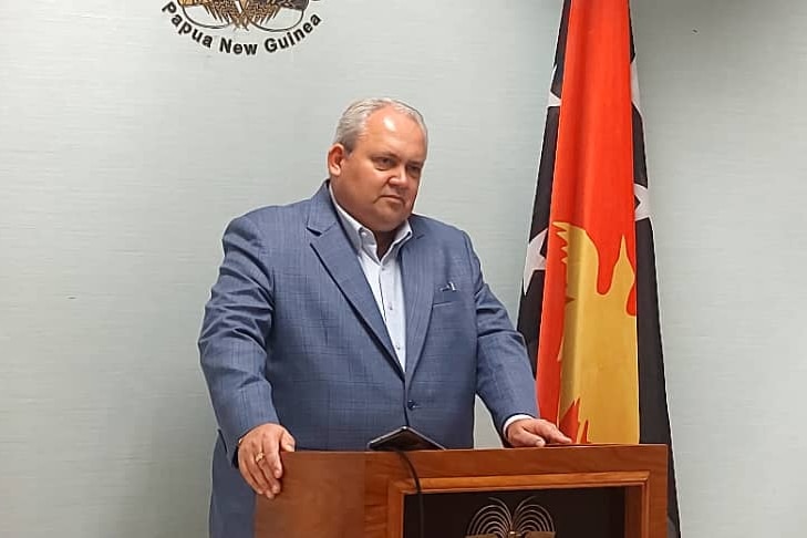 A plump man in a grey suit stand between two PNG flags as he speak from behind.