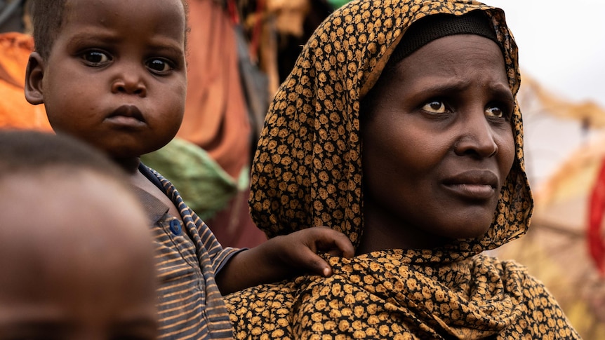 A woman wearing a yellow and black head garment cradles a sleeping baby while a toddler places his hand on her shoulder