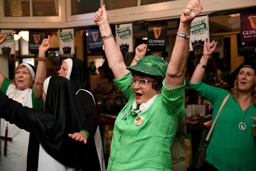An older woman celebrates with arms in the air in green costume