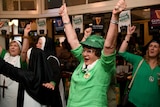 An older woman celebrates with arms in the air in green costume