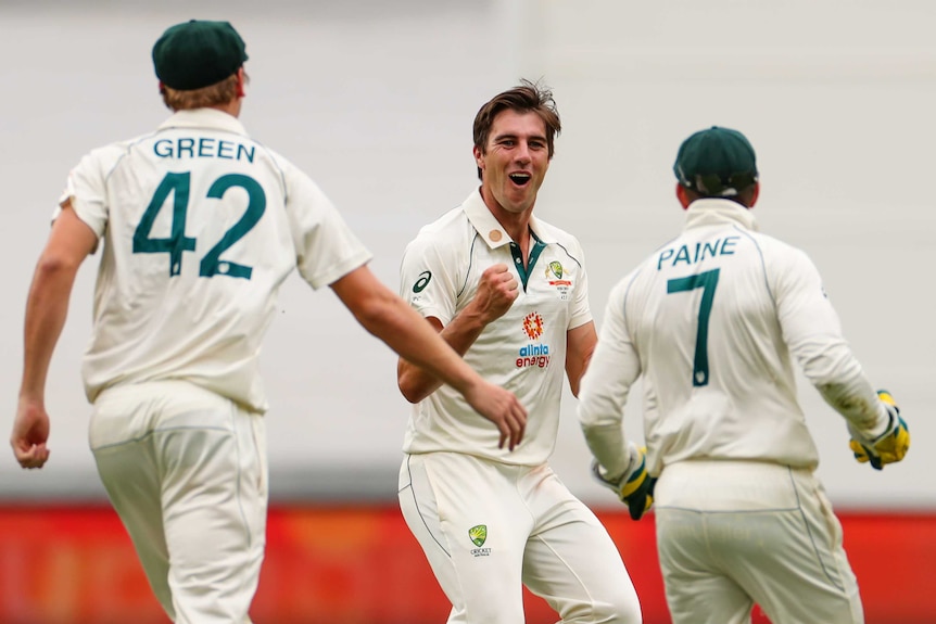 Pat Cummins runs with his fist clenched as two men wearing cricket whites run towards him