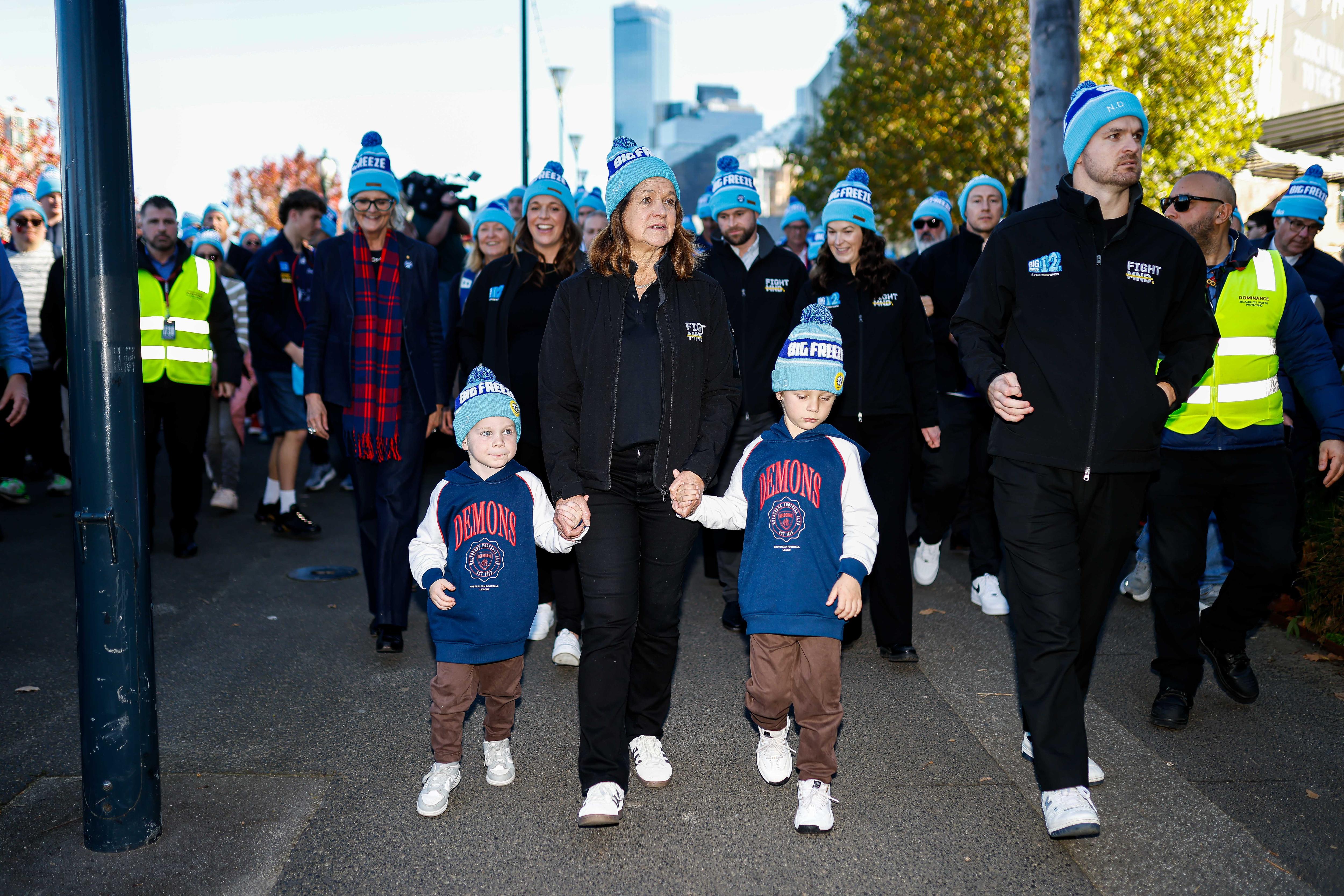 A woman dressed in black with a blue and white beanie holds hands with two children, leading a crowd in the same beanies.