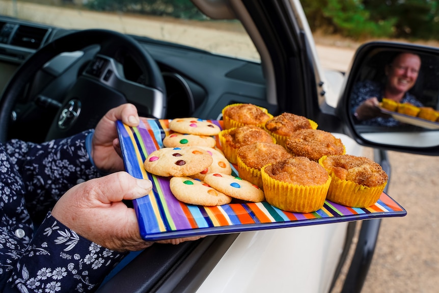 A woman sitting in a car holds a colourful plate decked out with cookies and muffins out of the driver window.