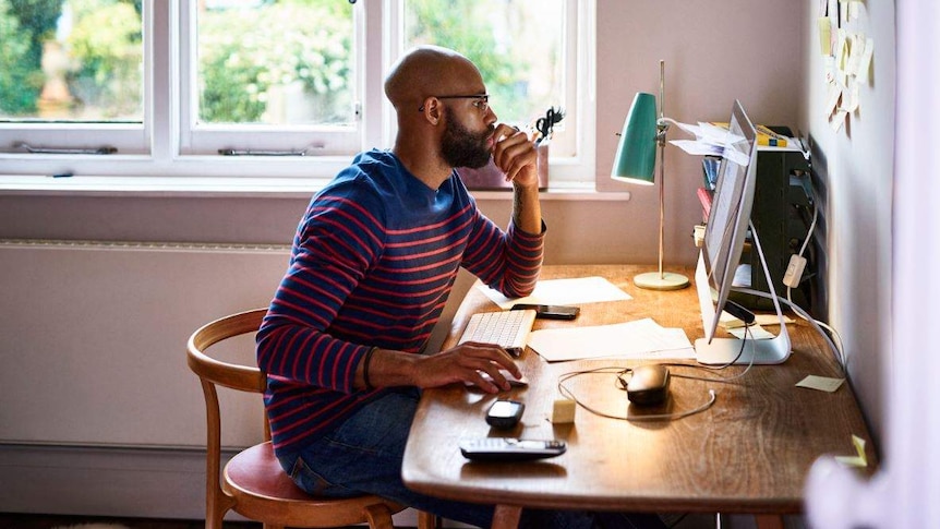 In a bright room, a man in striped jumper sits at a desk looking at a computer screen, with hand to mouth as if in thought.