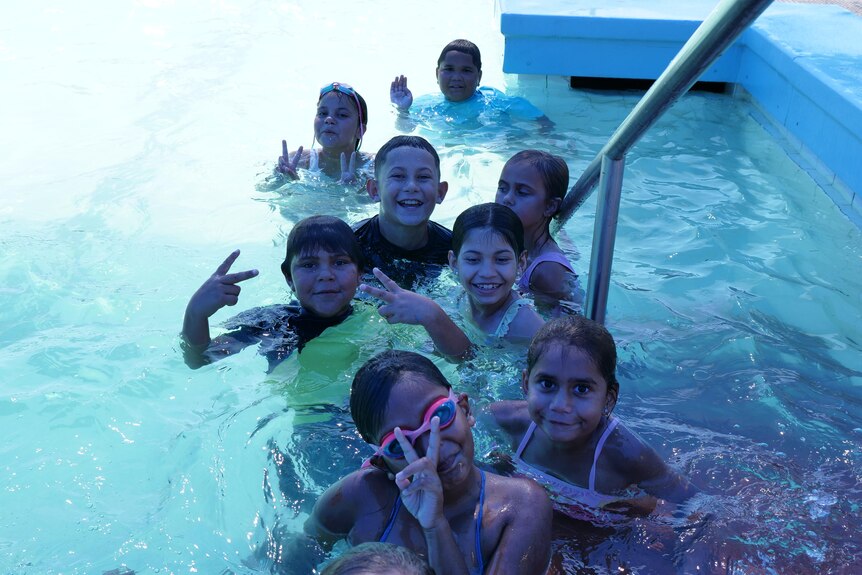 A group of kids splashing in the pool, looking at camera