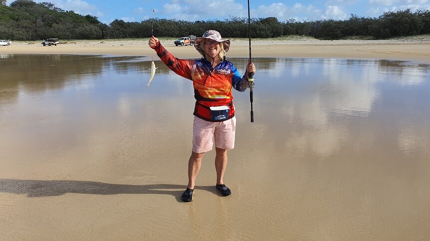 A woman stands on a beach holding a fish she has caught and a fishing rod