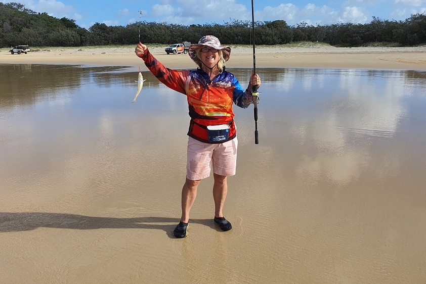 A woman stands on a beach holding a fish she has caught and a fishing rod