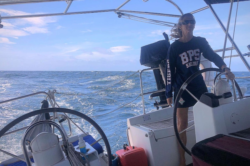 Woman helms a yacht by port steering wheel on port tack in open sea. Windy. Camera looking astern.