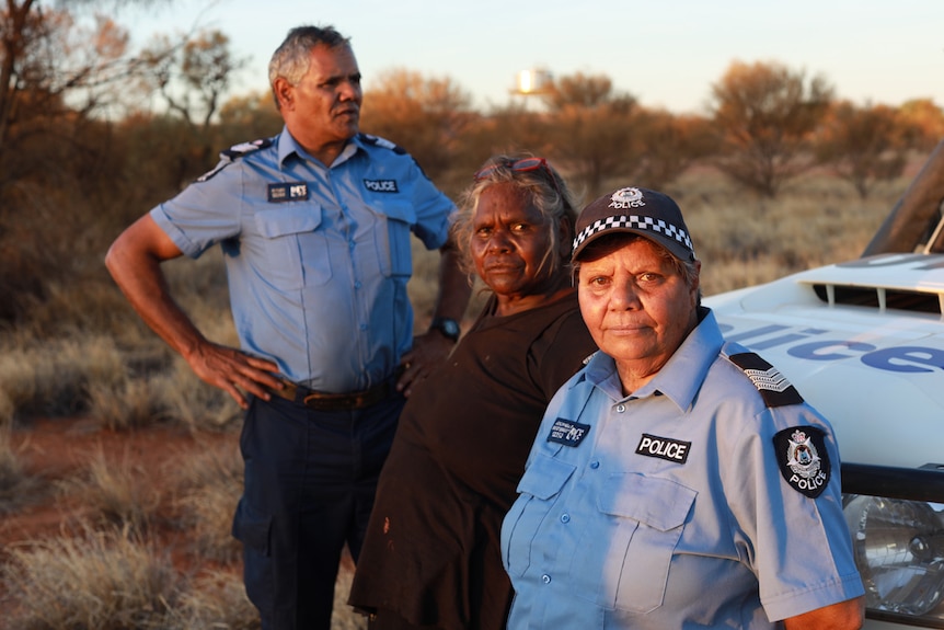 Three Indigenous Australians, two in police uniforms and one in black clothing, standing in front of a police car in the dessert