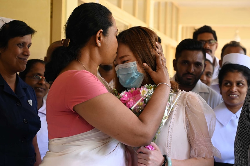 Woman in white sari and pink blouse kisses masked woman on forehead in front of onlookers.