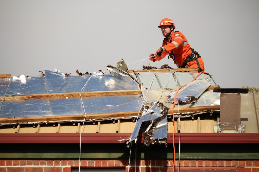 A man in orange overalls uses a rope to tie down sarking that is underneath what is a missing roof