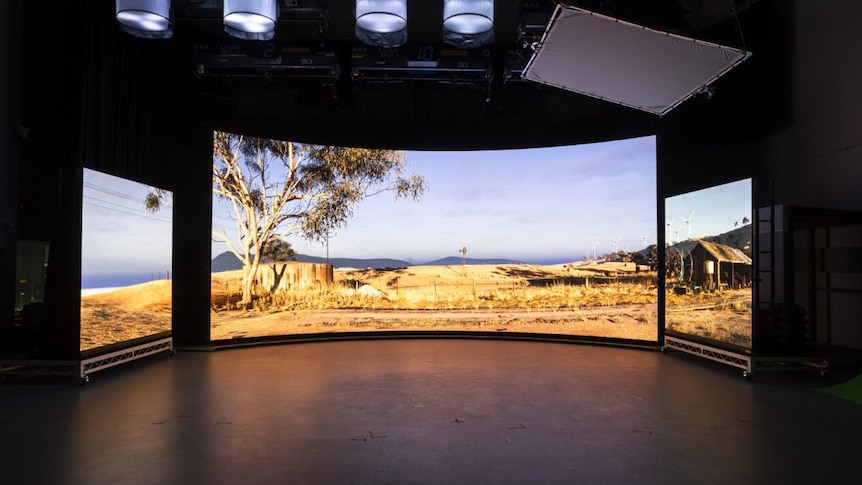 Three large screens showa scene of a tree and paddock in the outback, within a black studio.