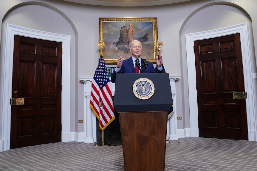 Joe Biden speaks at a lectern in front of a fireplace and US flags in a carpeted room.