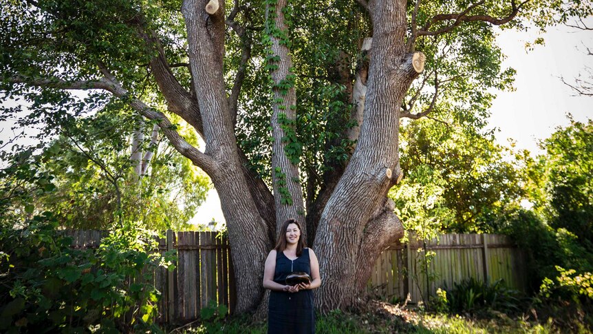 Miriam standing under a huge tree in backyard, holding small metal drum.