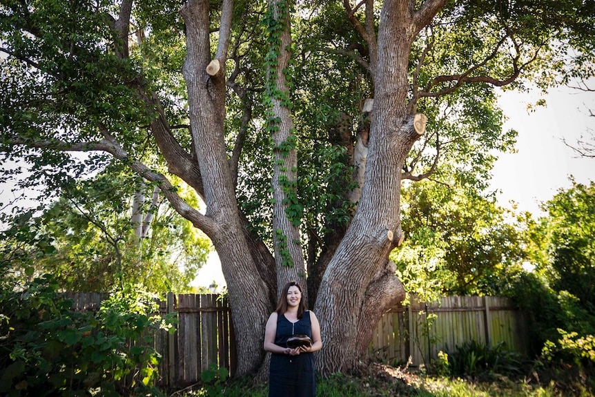 Miriam standing under a huge tree in backyard, holding small metal drum.