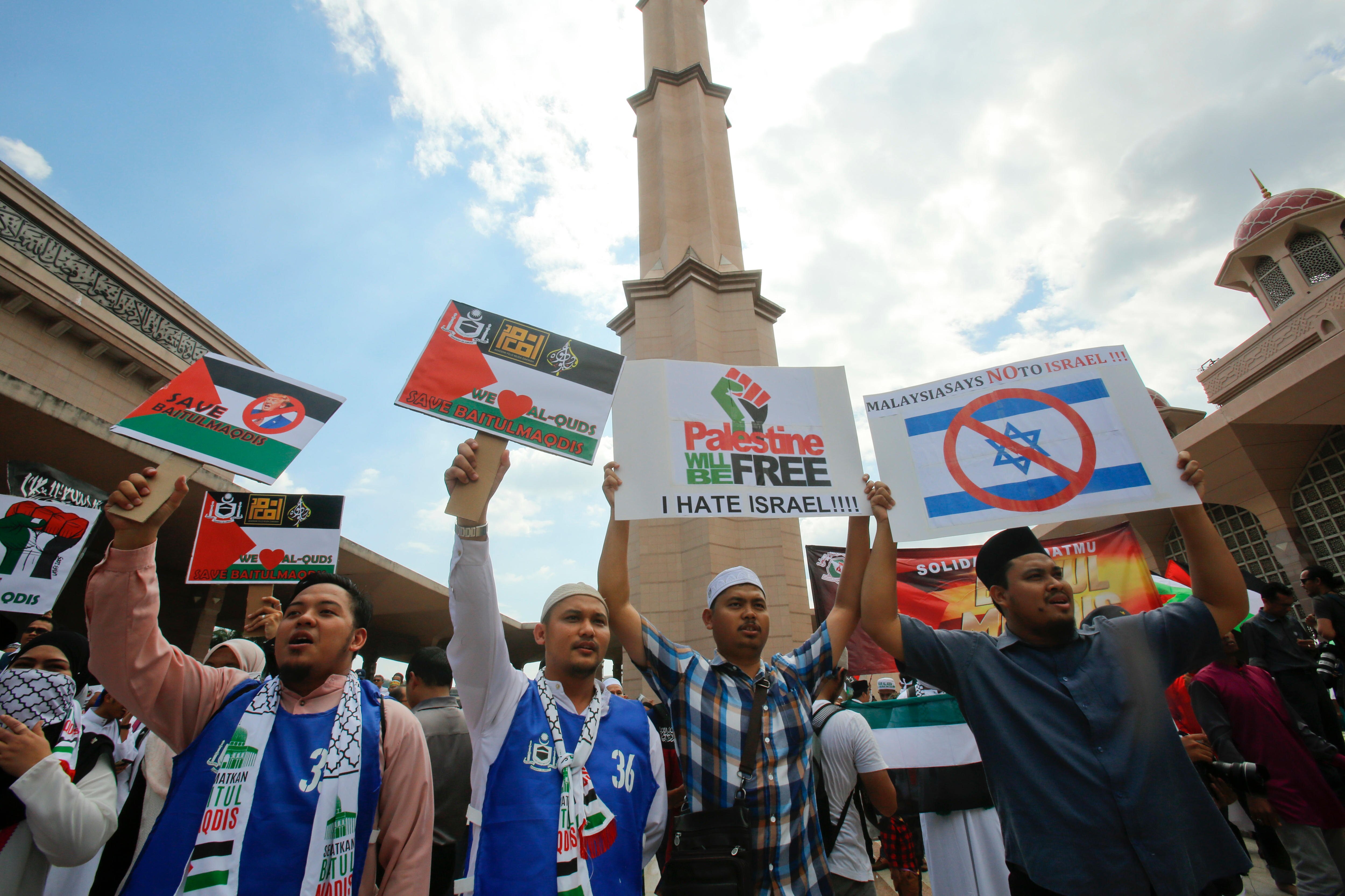 4 men with white, green, red and black scarves around their neck hold up placards of the Palestinian flag in front of a mosque