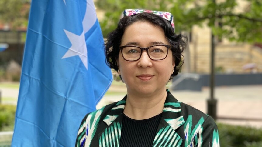 A woman with short dark curly hair stands wearing a green and black blazer next to a blue and white East Turkestan flag.