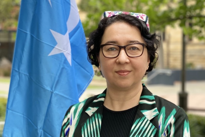 A woman with short dark curly hair stands wearing a green and black blazer next to a blue and white East Turkestan flag.