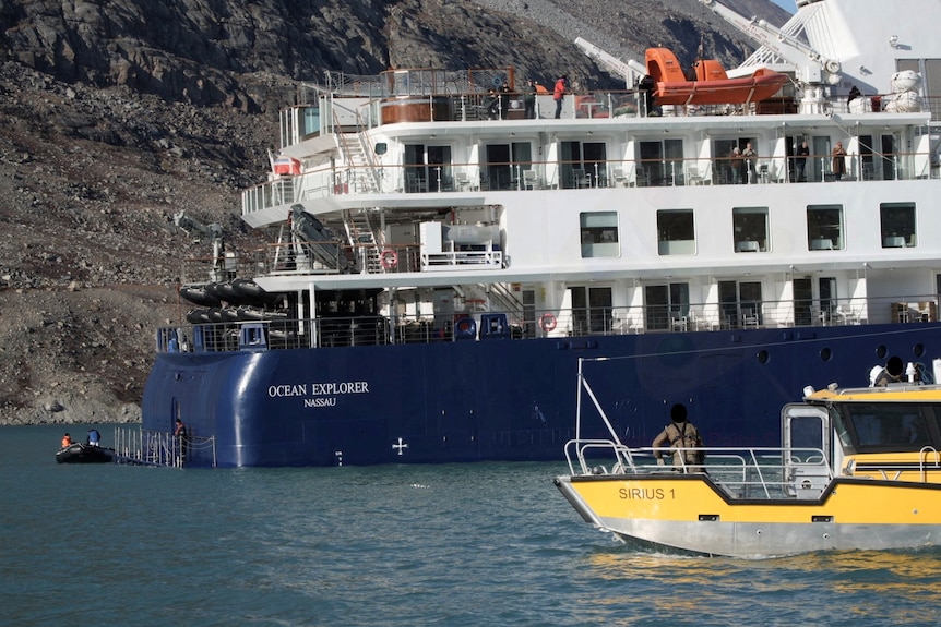 A cruiseship is stuck in low water, two smaller boats have come up to it, a rugged hillside is in the background