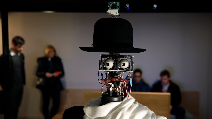 A close-up of humanoid robot with fake eyes and lips, wears a hat and a camera on its head, and a throw around its neck