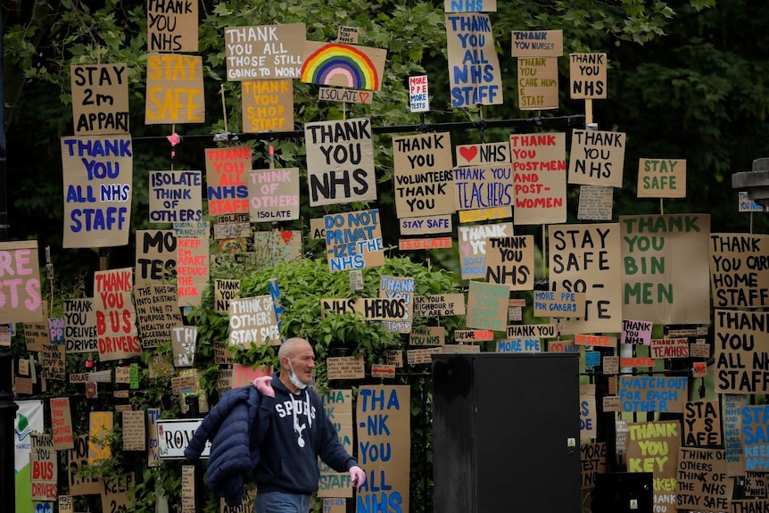 A man wearing a mask under his chin walks past cardboard signs with slogans like 'Thank you NHS' and 'Stay safe' on them.