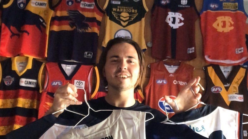 A man smiles while holding black and white Carlton jumpers in front of a wall of different jumpers.