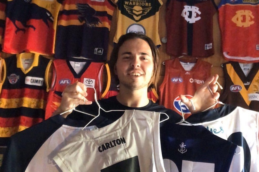 A man smiles while holding black and white Carlton jumpers in front of a wall of different jumpers