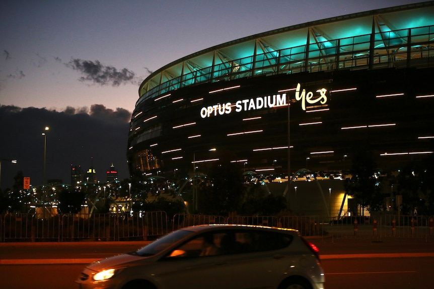 Perth Stadium with some blue and yellow lights on it as a car drives past the road in front and the city skyline is in the back