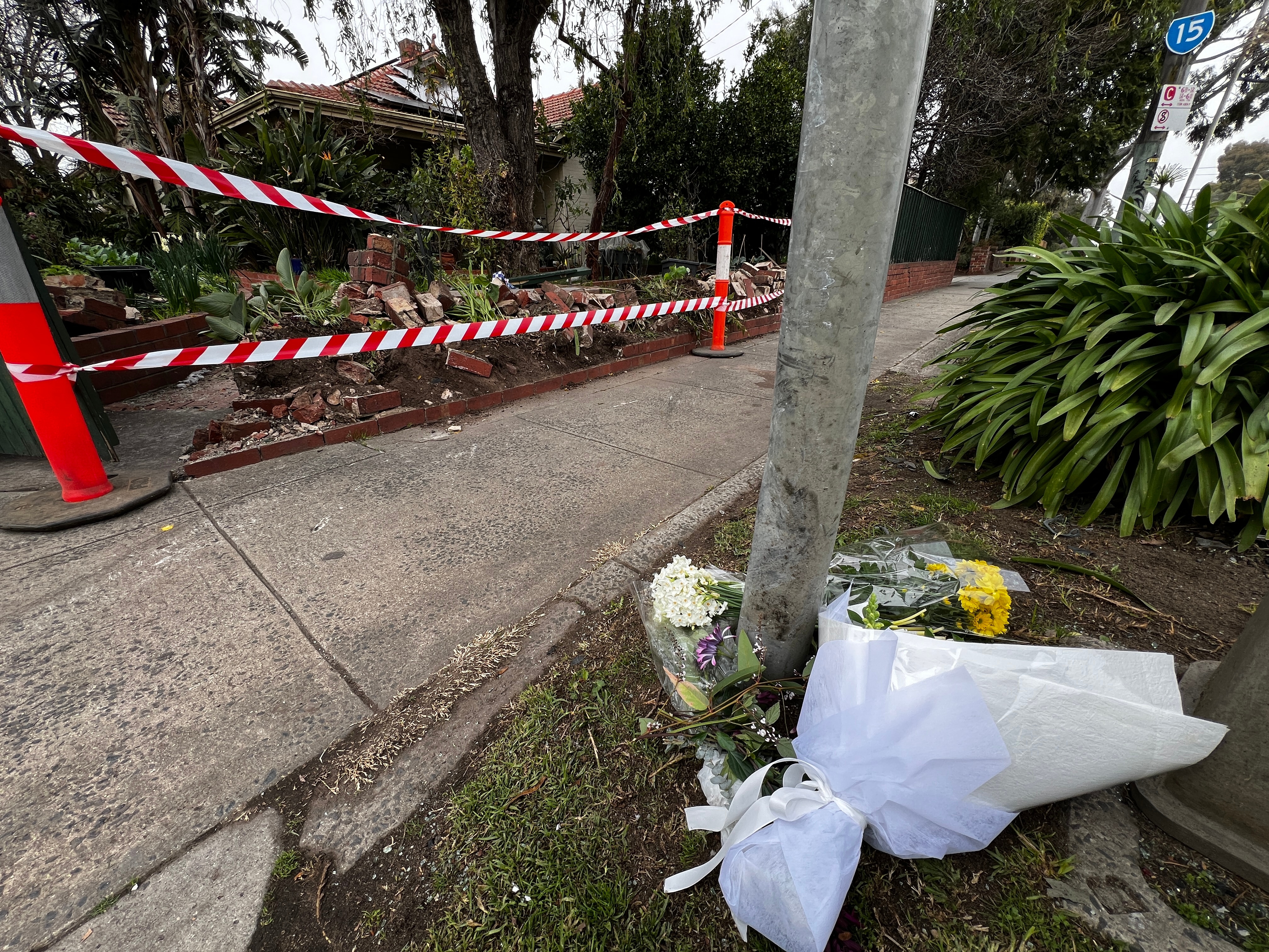 Bouquets of flowers lie on the pavement beside emergency services tape over a crushed brick fence.